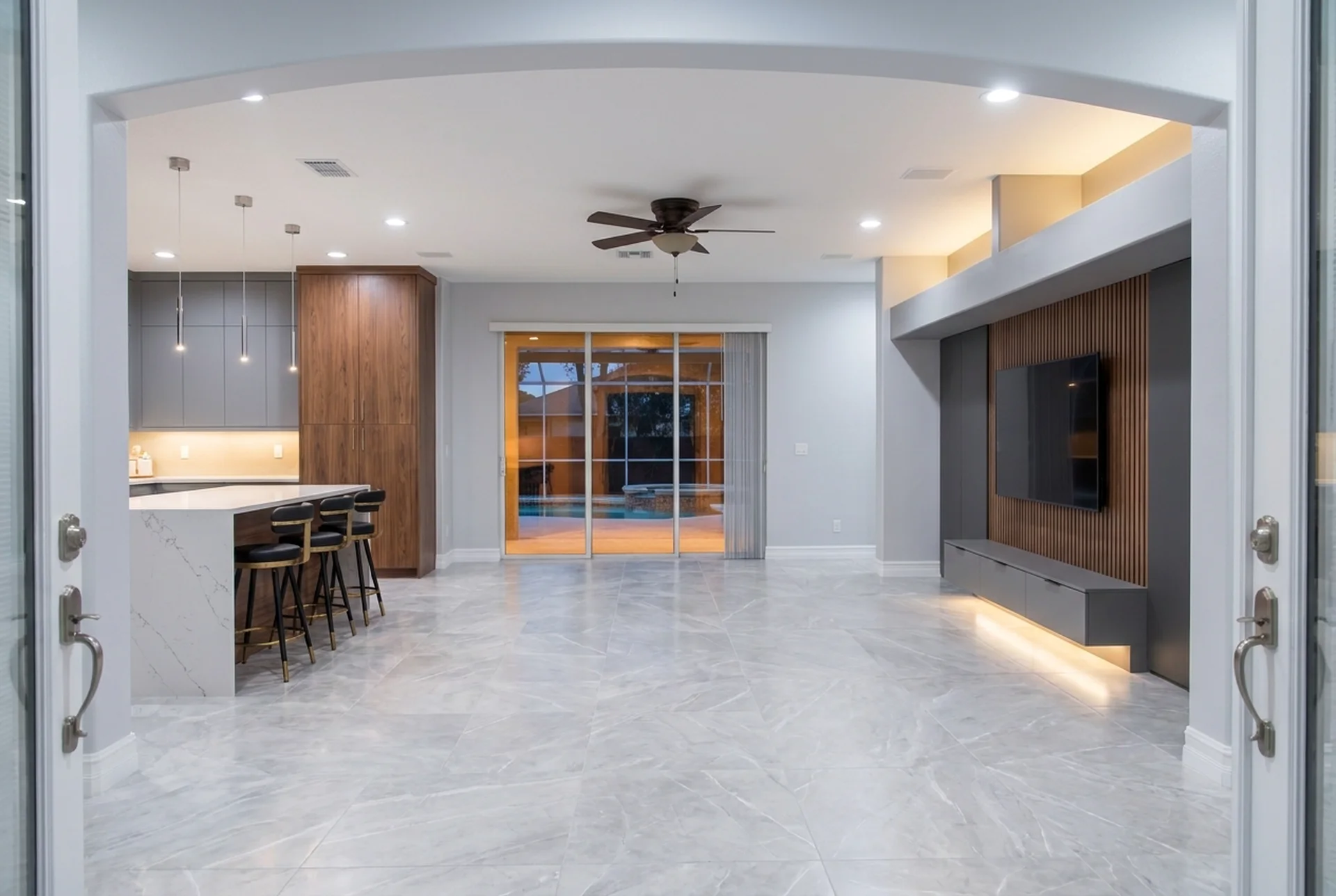 AI-assisted interior renovation concept: the view from inside the front doors — media wall right, kitchen left, and the lit pool through the sliding doors ahead.